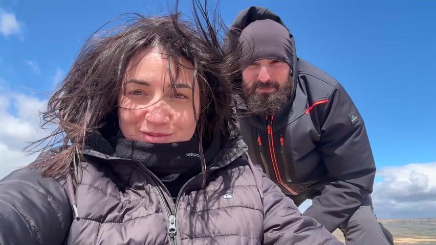 A woman and a man are standing outside on a windy day. The woman is in the foreground, holding the camera and looking at it with a slight smile. Her hair is being blown around by the wind. The man is standing behind her and to her right, also smiling at the camera. He is wearing a beanie and has a beard. Both are wearing dark colored jackets. The sky is blue with some wispy clouds. In the distance, the landscape is arid and brown.