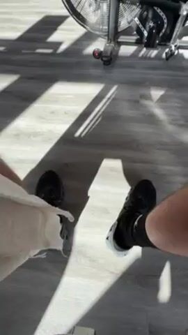 The video presents a first-person perspective of a person's legs and feet on a gray, wood-like floor in a brightly lit room. The person wears black athletic shoes and socks. The left leg appears to be wrapped in a white bandage or towel. In the background, part of an exercise bike is visible. Sunlight streams in through a window, creating shadows on the floor. The camera is stationary.