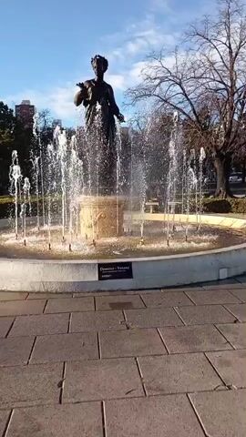 This media file shows a bronze statue of a woman standing in the center of a circular fountain. The statue is positioned on a pedestal, and water sprays from multiple jets surrounding it, creating a dynamic effect. The fountain itself is set within a paved area, with a white concrete rim. A dark plaque with white text is visible on the rim of the fountain, which reads "Deméter - Verano".

In the background, a line of trees with bare branches suggests a cooler season, possibly late autumn or winter. Behind the trees, modern buildings rise into the sky, indicating an urban setting. The sky is blue with scattered clouds, and the sunlight suggests it is daytime, likely late morning or early afternoon given the angle of the light.

The overall scene is tranquil and picturesque, capturing a moment of public art and nature in an urban environment. The activity is simply the operation of the fountain, with the statue serving as its centerpiece. The location cues, such as the architecture and the plaque text (possibly indicating "Demeter - Summer" in Spanish or Italian, given the context of La Plata), help place the scene in La Plata, Argentina.