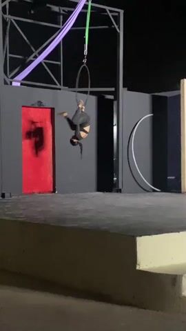 A woman in a black leotard and pants is performing aerial acrobatics on a metal ring suspended from a frame. The ring is hanging from a green strap, and there is a purple silk fabric also hanging from the frame. The woman is upside down, with one leg extended upward and the other bent. The performance is taking place on a stage at night. The stage has a gray surface and a backdrop of black walls. There is a red door on one wall and a white circle on another. The camera is stationary.