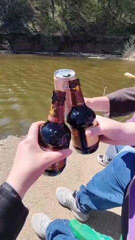 The video begins with a close-up shot of three arms holding two brown glass bottles and a pink can against a river backdrop, with the people wearing casual clothing and shoes. The hands holding the bottles clink them together, while the can is nearby but not clinked. The camera then pans to show the river, reflecting sunlight, surrounded by trees under a clear blue sky. The water appears to transition from a murky brown to a darker blue as the camera pans.