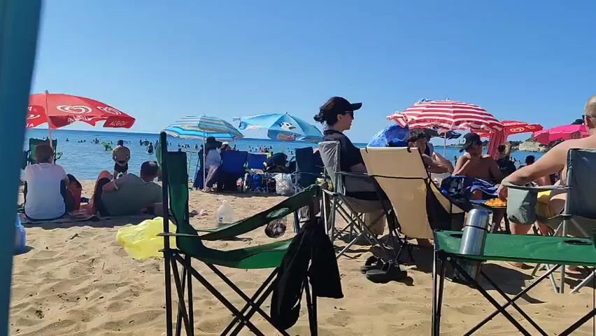 The video shows a crowded beach on a sunny day. People are sitting in beach chairs and under umbrellas. Some people are in the water, while others are relaxing on the sand. The sand is light brown, and the water is blue. The sky is clear and blue. There are several beach umbrellas, some red and white striped, others blue, and one red umbrella with a white logo. In the foreground, there are several folding chairs. A yellow plastic bag is visible near one of the chairs. A group of people are seated at a table on the beach, some with drinks and snacks. The people are dressed in casual summer clothing.