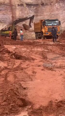 An excavator is loading a dump truck with dirt. Three men are standing near the excavator, and one man is standing near the dump truck. The ground is covered in red dirt, and there are tire tracks in the dirt. The scene appears to be a construction site or a mine.