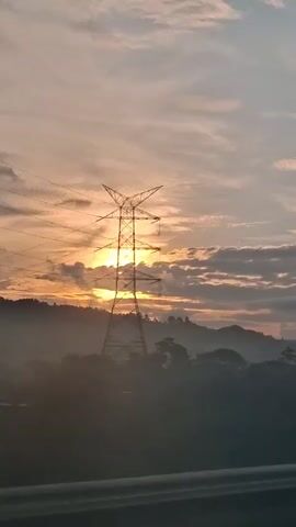 A sunrise is seen over a hilly, forested landscape. The sun is partially obscured by clouds and a large electricity pylon is in the foreground. The sky is a mix of pink, orange, and light blue, with some scattered clouds. The hills are covered in trees, and some mist can be seen in the valleys.