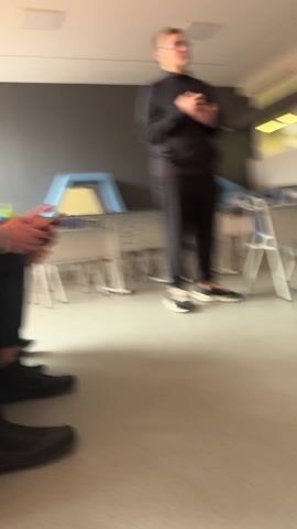 The video depicts a room with three people sitting on a bench and two people standing. The first person sitting is using a smartphone, and the person next to them looks on. The person standing closest to the camera wears black athletic clothing, including a jacket. The second person standing wears a brown jacket, a striped shirt, and blue jeans. They have their hand on their hip and appear to be speaking. Tables are stacked along the back wall, and the room appears to be in a school or office setting.