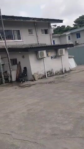 The video shows an outdoor paved area in front of a two-story building with a light gray facade. There are several air conditioning units mounted on the side of the building. A black SUV is parked to the right of the frame, and a person is partially visible near a wooden post on the left side of the frame. The sky is overcast. The camera appears to be moving, showing different angles of the building and the SUV.