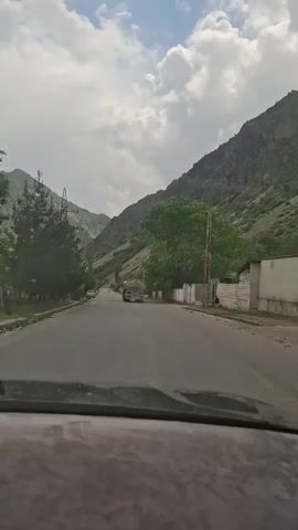 A car drives down a narrow road flanked by mountains under a cloudy sky. A large tanker truck drives in the opposite direction. A parked silver SUV is visible to the left. Trees line parts of the road, and a power line tower stands on the left side. Buildings and a person are visible on the right side of the road.
