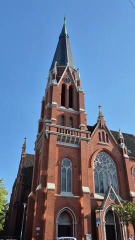The video shows the facade of a red brick church with a tall, dark spire against a clear blue sky. The church has several arched windows, including a large rose window above the entrance. A clock is visible on the spire. In front of the church, there are a few cars parked on the street, and a person can be seen standing near the entrance. The camera is stationary and captures the church from a low angle, emphasizing its height. There is no discernible audio in the video.