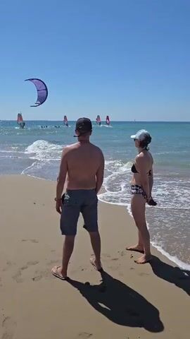 The video shows a crowded beach on a sunny day. There are many people walking and sitting on the sand. In the water, several people are windsurfing and kitesurfing, with their colorful sails visible against the blue sky and ocean. The waves are gentle, and the water appears clear. The overall atmosphere is one of leisure and recreation.