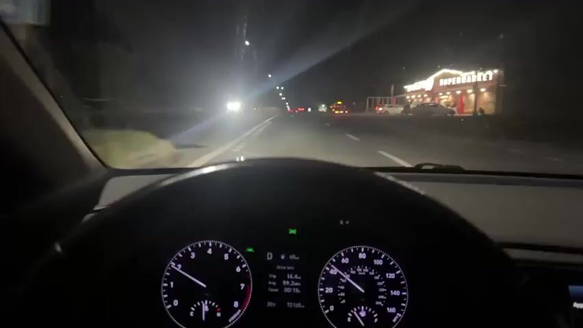 This is a first-person perspective video recorded from inside a car at night. The dashboard is clearly visible, showing the speedometer, tachometer, and trip computer. The speedometer indicates a speed of approximately 65 km/h. The trip computer displays trip distance of 14.4 km, average speed of 59.2 km/h, and trip time of 00:15. The current temperature is 20°C and the odometer reads 72165.

The scene outside the car is a road at night, illuminated by streetlights and the headlights of oncoming vehicles. To the right, there's a building with a sign that appears to say "SUPERMARKET" with lights on. There are other cars on the road and parked near the supermarket. Trees and foliage are visible on the sides of the road, suggesting a semi-urban or rural setting. The overall impression is that of a nighttime drive on a relatively well-lit road, possibly on the outskirts of a town. The location is identified as Metsamor, Armenia.