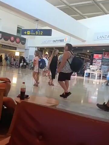 The video shows an indoor scene, likely an airport terminal, given the visible "LANZAROTE DUTYFREE" sign and a gate sign indicating "Terminal Puertas / Gates 9-12". Numerous people are seen walking through the bright, spacious area with a light-colored, reflective floor.

In the midground, a young girl in white shorts and a pink top walks past, followed by a woman in a black skirt and patterned top with a backpack. A man in a black t-shirt and shorts, carrying a blue backpack, walks in the same direction. Another man in a light blue shirt and shorts, and a woman in a long black dress are also visible, moving amongst other scattered individuals in the background.

In the foreground, partially visible, is a dark red, tufted armchair and a wooden chair. On a small, round wooden table between these chairs, a red Coca-Cola bottle is placed. The overall atmosphere appears calm and routine for an airport setting, with people either passing through or waiting. Other partially visible signs include "Cajas" and "SMOKING AREA". The bright lighting suggests it is daytime. The location is consistent with Lanzarote Airport in San Bartolomé, Spain.