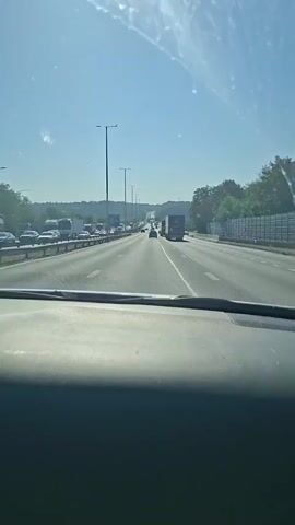 The video shows a view from inside a car driving on a multi-lane highway on a clear, sunny day. The car's dashboard is visible in the foreground. The highway has multiple lanes of traffic with cars and trucks moving in the same direction. Streetlights are visible along the side of the highway. Trees and some structures are visible beyond the highway. There is some light haze or dust on the windshield. The overall impression is a typical highway driving scene during daylight hours.