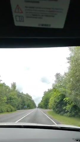 The video shows a view from inside a car driving on a paved road bordered by lush green trees on both sides. The sky is overcast with clouds. Raindrops are visible on the windshield and the windshield wipers are operating. There are a few cars visible on the road ahead. A warning label is affixed to the interior top of the windshield.
