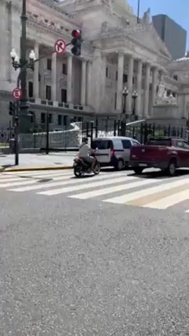 The video shows a street in a city with European-style architecture. The buildings are made of light-colored stone and have classical architectural details like columns and domes. There are several cars and a motorcycle on the road. The camera pans from right to left, following the flow of traffic. The weather appears sunny with some clouds in the sky. There are zebra stripes on the crosswalk. There is a no entry sign and a traffic light on the corner of the street.