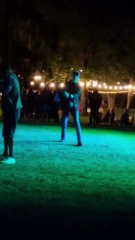 The video shows a nighttime outdoor gathering. The ground is covered in green grass, illuminated by green and blue lights. In the background, trees are visible, adorned with strings of warm white lights. Several people are scattered throughout the scene, some sitting and some standing. In the foreground, two individuals are clearly visible. The person on the left is wearing a green top and dark shorts, with white shoes. The person on the right is wearing a dark jacket and blue jeans, with a baseball cap. This person is in motion, appearing to be dancing or gesturing. The overall atmosphere suggests a casual event or party.