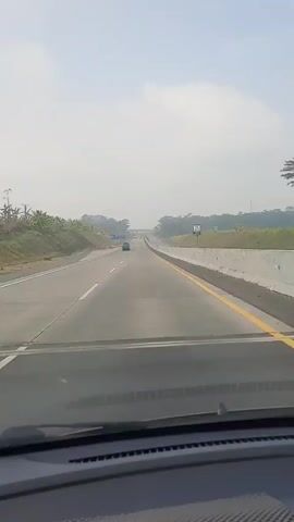 The video is a first-person perspective from inside a car driving on a highway. The road is paved and appears to be in good condition, with clear lane markings. There are guardrails on the right side of the road and a slight incline of land with vegetation on the left. The sky is overcast with a hazy appearance. There are a few other cars visible on the highway in the distance, all traveling in the same direction as the camera car. A sign with blue lettering is visible on the left side of the road. The dashboard of the car is visible at the bottom of the frame.