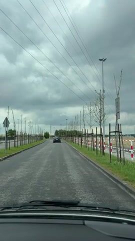 The video is shot from inside a vehicle moving down a paved road, and shows a view of a theme park on the right side of the road, marked by a fence. The theme park features at least one roller coaster with loops and a structure resembling a castle or turret. Small, newly planted trees line the road, supported by stakes. Yellow construction vehicles are visible within the theme park. The sky is overcast with heavy clouds. A black car is visible in the distance moving in the same direction. Power lines stretch across the sky above the road. The road surface is dark and slightly wet, possibly from rain.