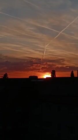 The video displays a picturesque sunset over Lahr/Schwarzwald, Germany. The sky is dominated by vibrant hues of orange and red near the horizon, gradually fading into lighter oranges, blues, and grays higher up. Several clouds are present, some catching the warm glow of the setting sun, while others appear as darker patches against the colorful sky. Distinct white contrails, likely from aircraft, crisscross the upper part of the frame, adding linear elements to the natural cloud formations. The sun itself is a bright, intense orb, partially obscured by the distant horizon and silhouetted structures. The lower portion of the video features a dark, silhouetted landscape composed of buildings and trees, standing starkly against the illuminated sky. No people or visible text are present. The scene evokes a sense of tranquil beauty at the close of day.