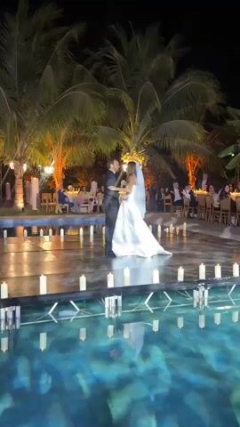A couple is slow-dancing at night in front of an audience. The bride is wearing a white dress and a veil. The groom is wearing a black suit. They are on a low platform above a pool, and lit candles line the platform. Tall palm trees are behind the couple, with lights illuminating them. The audience members are seated at tables on either side of the dance floor.