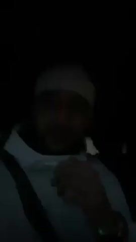 A man is sitting in a car in the dark. He is wearing a white hoodie, a white beanie, and a seatbelt. He is talking to the camera, but his face is not very well lit. There is music playing in the background.