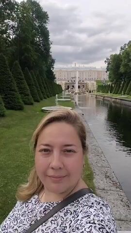 A woman takes a selfie in front of a canal with fountains and a large building in the background. The woman is smiling and looking at the camera. The canal is lined with trees and manicured bushes. The building in the background appears to be a palace or a grand house. The sky is cloudy.