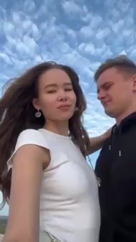 A young couple is standing together outdoors. The woman has long, wavy, brown hair, and is wearing a white t-shirt. The man has short, light-brown hair, and is wearing a black hoodie. They kiss briefly, then smile at the camera. The background is a blue sky with fluffy white clouds. The camera pans slightly to show a sandy landscape, possibly a beach or desert, with water visible in the distance.