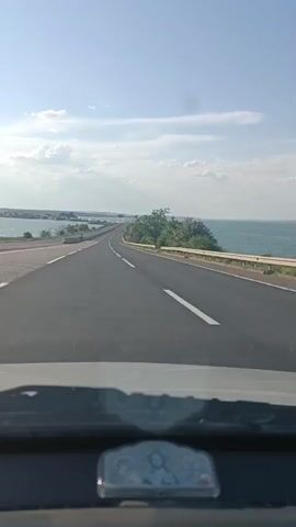 This media file captures a view from inside a vehicle traveling on a paved road. The road curves gently to the right and appears to be a multi-lane highway, indicated by the white lane markings. On the right side of the road, a metal guardrail runs alongside a body of water that stretches to the horizon. The water is a deep blue, suggesting it is a sea or a large lake.

The landscape is predominantly open, with sparse greenery along the roadside. In the distance, a low-lying landmass with trees is visible, and further on, a long bridge or causeway spans across the water. The sky is a bright, clear blue with scattered white clouds, indicative of fair weather. The sun appears to be high in the sky, suggesting it is daytime, likely late morning or early afternoon.

The perspective is from the driver's seat, with the dashboard and a small, reflective object on it visible in the foreground. The object on the dashboard appears to be a digital clock or a similar gauge, but the details are too blurry to discern specific text or numbers.

The scene conveys a sense of travel and open road, with the vastness of the water and sky creating a peaceful and expansive atmosphere. There are no people clearly visible in the media, but the presence of the road and the guardrail suggest a developed area, possibly a coastal highway. The general setting suggests a location near a large body of water, consistent with the provided location context of Altestove, Ukraine, which is known for its coastal features.