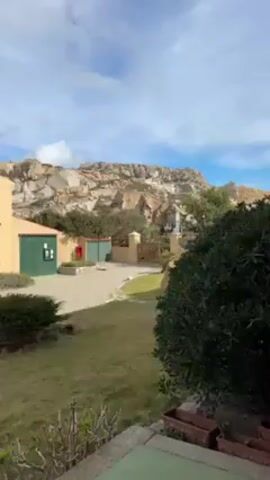 The video shows a view of a light yellow, two-story building with green window shutters and a red tile roof. There is a grassy lawn in front of the building, with a stone driveway leading to a gate. Beyond the gate, there is a rugged, rocky mountain range under a blue sky with white clouds. A black dog walks across the lawn.
