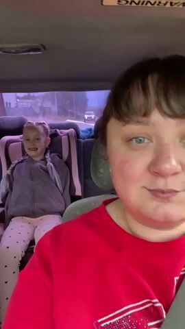 This is a selfie video taken from inside a car. A woman with brown hair and a red shirt is in the foreground, smiling. In the background, a young girl with blonde braided hair and a grey hoodie is sitting in a car seat. The car appears to be in motion, as indicated by the blurred background of trees and streetlights. There is a warning label on the car's ceiling.