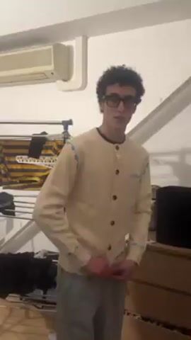 The video shows two young men in a bedroom or dressing room. The first man, with curly dark hair and glasses, is wearing a light tan button-down sweater with light blue details and gray sweatpants. He makes a peace sign with his hand at the start. He then turns as another man walks into frame. The second man has curly dark hair and wears a dark brown button-down sweater with pink details over a white undershirt. Both men look at the camera and smile. The first man then bends down out of frame. The background shows clothing racks, a dresser, and a small air conditioning unit.