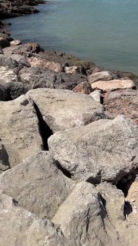 The video shows a body of water and a rocky shoreline. In the background, there is a town with buildings and a beach. The water is a light blue-green color. The sky is clear and blue. There are no people visible in the foreground or midground. The focus is on the rocks and the water. The camera is moving slowly, panning across the scene.