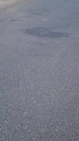 The video shows an asphalt road with cars parked on the left side. The cars appear to be SUVs and sedans. The sun is shining, casting shadows on the road. The environment appears to be an urban or suburban setting, possibly a parking lot or a street. The video is shot from a low angle, looking forward.