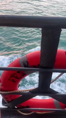 The video opens with a close-up of a red life preserver ring, secured by a rope and attached to a dark metal railing. The railing is likely part of a boat or ferry. The background reveals the ocean, with gentle waves and hints of white foam.

As the camera pans, the focus shifts to the horizon. The sun is setting, casting a warm, orange glow across the sky and reflecting on the water's surface. Distant islands, silhouetted against the vibrant sunset, are visible on the horizon. The sky is partly cloudy, with some darker clouds contributing to the dramatic effect of the sunset. The overall scene suggests a tranquil and picturesque moment at sea.

There is no visible text in the media file. The time of day is clearly sunset. The location context of Thong Nian, Thailand, aligns with the visual cues of islands and the tropical sunset. The emotions conveyed are peaceful and serene, characteristic of a sunset over the ocean. The activity taking place is a journey by boat, indicated by the railing and the life preserver.