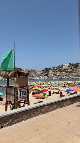 The video opens to a beach scene under a clear blue sky. The beach is populated with beachgoers relaxing under colorful umbrellas, most of which are red, yellow, orange, pink, and blue. The sand is light brown, and there are rocks and a stone wall separating the beach from a walkway. Beyond the beach, there is a body of water filled with numerous boats and yachts. In the background, a picturesque town with buildings is nestled on a hillside, surrounded by lush green trees. The camera pans slightly to the right, revealing more of the harbor and the town, with a few people visible on the beach. Towards the end of the video, the perspective shifts, showing a glimpse of a palm tree in the foreground. The overall atmosphere is one of a sunny day at a popular coastal destination.