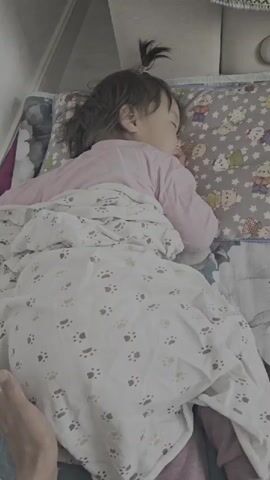 A high-angle medium shot shows a young child sleeping on their side, facing away from the camera. The child is wearing a pink long-sleeved shirt and is covered by a white blanket adorned with paw prints. A pillow with cartoon characters is positioned under the child's head. A person's hand with a ring is visible in the lower left corner of the frame, resting on the blanket near the child's legs. The background is out of focus, but appears to be an indoor setting with furniture. The lighting is soft and diffused.