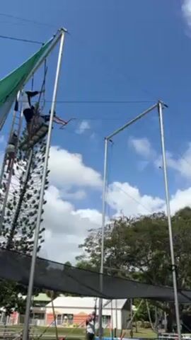 The video shows a person performing on a flying trapeze. The trapeze is set up outdoors, with trees and buildings visible in the background. The person swings on the trapeze. There are other people present, some on the platform of the trapeze structure and one walking below. The sky is blue with some clouds.