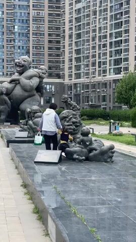 The video shows a scene in what appears to be a park or public space, set against the backdrop of tall apartment buildings. A woman, dressed in a white vest and dark pants, stands with a young child near a series of large, somewhat abstract sculptures. The sculptures are gray and depict figures, some of which appear to be laughing or smiling. The woman appears to be helping or supervising the child as it interacts with the sculptures, possibly climbing on them. The surface beneath the sculptures is a dark, shiny material that seems to be slightly wet. There are also pigeons visible in the background.
