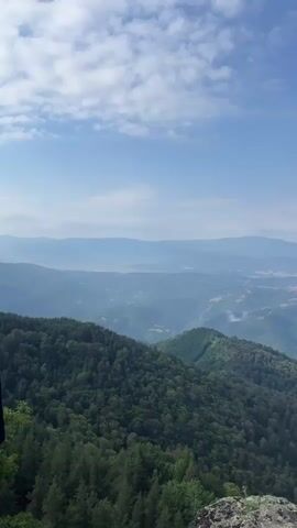 The video is a vertical shot from a high vantage point, looking out over a vast, hazy mountain landscape. The foreground features a rocky outcrop and the tops of evergreen trees. Rolling, densely forested hills dominate the middle ground, with a lighter green, more open area visible on one slope. In the distance, layers of mountains fade into a pale blue haze, and a light-colored settlement or city is visible nestled within a valley. The sky above is a clear blue with scattered, wispy white clouds. The overall impression is one of expansive natural beauty and distance.