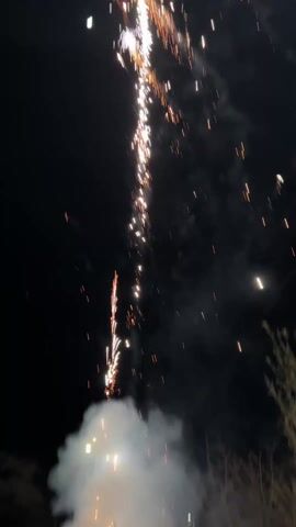 The video shows a group of people holding a banner at night, with fireworks going off in the background and on the ground. The banner has writing on it. People are standing and holding the banner up, while fireworks explode in the sky and on the ground near them, creating a smoky atmosphere. A flashlight shines on the banner.  The ground is paved with stone blocks.