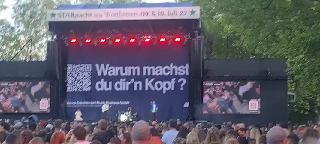 A crowd of people watch a performance on an outdoor stage at dusk. The stage is set up with a large black curtain backdrop displaying a QR code and the German text "Warum machst du dir'n Kopf?". Three musicians are visible on stage, one playing a guitar, another behind a drum set, and a third singing into a microphone. Two large screens on either side of the stage also show images of people holding up phones. The atmosphere suggests a music festival or concert.