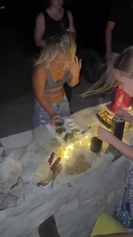 A group of people are gathered around a low stone structure at night. A woman with blonde hair in a gray crop top and ripped jeans is holding a small bowl and touching her hair. To her right, another person with blonde hair, wearing a black and white patterned top, is reaching towards a tray on the stone. The tray contains several small bowls with liquids and what appears to be food items. Next to the tray, there are string lights. A plate of food with what looks like hot dogs and bread is on the left side of the stone structure. Other people are partially visible in the background, and the ground appears to be sand.
