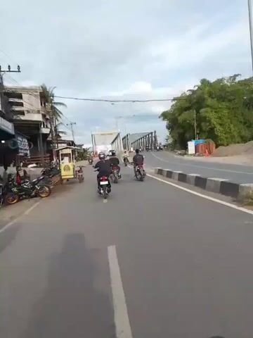 The video shows a road with several motorcycles and a bridge in the distance. On the left side of the road, there are buildings and parked motorcycles. A small food stall is also visible. Several people are riding motorcycles, moving towards the bridge. The sky is cloudy. The overall scene appears to be a busy street in a tropical or subtropical area.