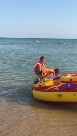 In this media file, a group of people are preparing for a water activity on a sunny day at Blagoveshchenskaya, Russia. The scene is set at the shoreline of a calm, clear body of water, likely the sea, with a sandy beach visible in the foreground.

The central object is a large, inflatable towable tube, primarily yellow and red, with the brand name "RIF" clearly visible. Several individuals are interacting with this tube, which is partially submerged in the water near the beach.

On the tube, a young girl wearing an orange life jacket and black swim bottoms is crouching, holding onto yellow handles. Beside her, a man in an orange life vest and green patterned shorts stands, appearing to assist or guide her. Further behind them, another person in a dark shirt and hat is also on the tube. On the right side of the tube, a person wearing a blue swimsuit and yellow life jacket is partially visible, reaching towards it.

In the background, the expansive ocean stretches to the horizon under a clear, bright blue sky, indicating it is daytime and the weather is fair. Several other people are visible on the water in the distance. Two individuals are on paddleboards, and one is on a jet ski. The jet ski, a white and silver model, is being operated by a person in dark shorts. The overall atmosphere appears relaxed and recreational, suggesting a day of summer fun and water sports.