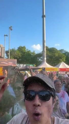 The video shows a person filming themselves at an outdoor celebration. The person is in a crowd of people, with their arms raised in the air. There is a lot of colorful powder in the air, suggesting the event is related to Holi or a color run. The person is wearing sunglasses and a hat, and their face and clothes are covered in powder. Other people in the crowd are also raising their hands and cheering. There are food vendors and tents set up around the event. The sky is blue and the sun is shining.