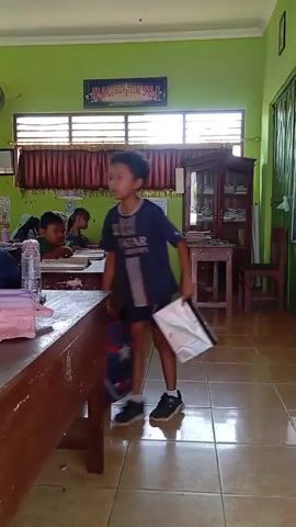 A young boy wearing a dark blue shirt walks across a classroom carrying a white and black bag in his right hand. He is walking away from the camera, and his face is not visible. The classroom has bright green walls, wooden desks with students sitting at them, and tiled flooring. There are windows with blinds and curtains at the back of the room, and a cabinet filled with books and other items is visible on the right side of the room. The video seems to be recorded from a low angle, with the camera positioned at desk height. The lighting is natural, coming from the windows.