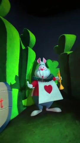 The video begins with a close-up of a character that resembles the White Rabbit from Alice in Wonderland, holding a trumpet. The scene transitions to a shot of a large playing card with a spade symbol, featuring a blurry, caricatured face. Next, a character that appears to be the Queen of Hearts, with large pink and white striped puff sleeves and a crown, is seen. This is followed by a closer view of the Queen of Hearts, identifiable by her red and black checkered dress and a distinctive crown. The video ends with a shot of people with their backs to the camera, one of whom is holding up a phone to record. The environment appears to be a dark, stylized setting with large, green, abstract shapes. A sign with red lettering that reads "Royal Croquet Game" is visible on the left side of the frame at the beginning of the video.