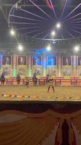 A circus performance is underway in an arena. The arena floor is covered in sand, with colorful flags lining the perimeter. In the background, a backdrop depicts classical Roman architecture with statues and murals. Above the arena, strings of lights create a circular pattern of illumination. In the center of the arena, a woman in a gladiator-style costume stands posed. Several horses with riders are positioned around the arena, some in motion. The lighting is theatrical, with spotlights and colored lights highlighting different areas of the performance space. The overall atmosphere suggests a reenactment of historical combat or a themed equestrian show.