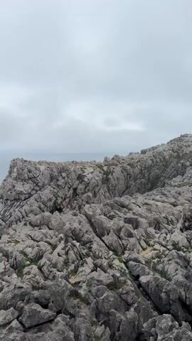 The video shows a panoramic view of a rocky coastline on an overcast day. The camera pans from right to left, revealing the rugged, gray rock formations of the cliff face covered in patches of green vegetation. The sea is a deep turquoise color, with whitecaps indicating choppy waters. In the distance, the horizon is blurred by mist or fog, and a lighter gray sky stretches across the frame. Further along the coast, other rock formations and cliffs are visible, jutting out into the sea. The overall impression is one of a wild and dramatic natural landscape.