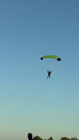 The video shows a person skydiving with a green and black parachute against a clear blue sky. The skydiver is descending and appears to be nearing the ground. In the foreground, there are several picnic tables and some people are seated at them. The environment appears to be an outdoor recreational area, possibly near an airfield. The skydiver is wearing a dark jumpsuit and a helmet. The parachute is open and inflated, providing lift and control for the descent. The overall scene depicts a skydiving activity with spectators observing from below.