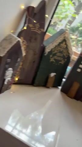 The video shows a windowsill decorated with small, house-shaped objects, string lights, and a vase of dried lavender. The houses appear to be made of wood and painted in various colors, with some featuring decorative elements like lace trim and gold lettering. One house has "Welcome Home" written on it. Branches with small butterflies and more string lights are arranged in the vase. Through the window, a garden scene is visible with trees, plants, and a hanging chair. The lighting suggests it might be daytime, possibly overcast. There are no people or animals visible in the video, and no actions or interactions are depicted. The camera pans slowly across the windowsill and then out the window.